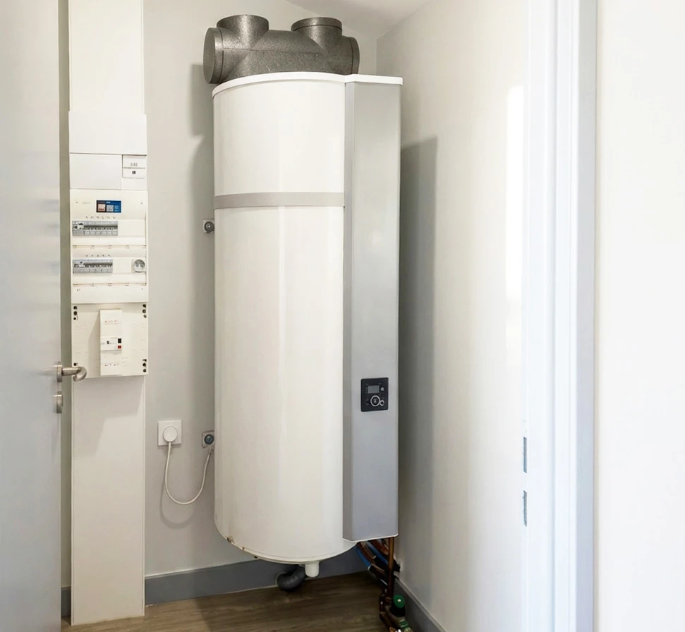 Photographie professionnelle d'une pompe à chaleur air-air moderne en cours d'installation par un technicien qualifié en gilet de chantier, fixée à la façade extérieure d'une maison contemporaine. L'unité extérieure est en acier inoxydable brossé, bien visible et clairement identifiable comme équipement de chauffage moderne. Le technicien porte des équipements de sécurité appropriés et manipule l'installation avec professionnalisme. La mise en place claire et professionnelle de ce système de chauffage moderne représente la performance énergétique et l'éligibilité aux aides de l'État.